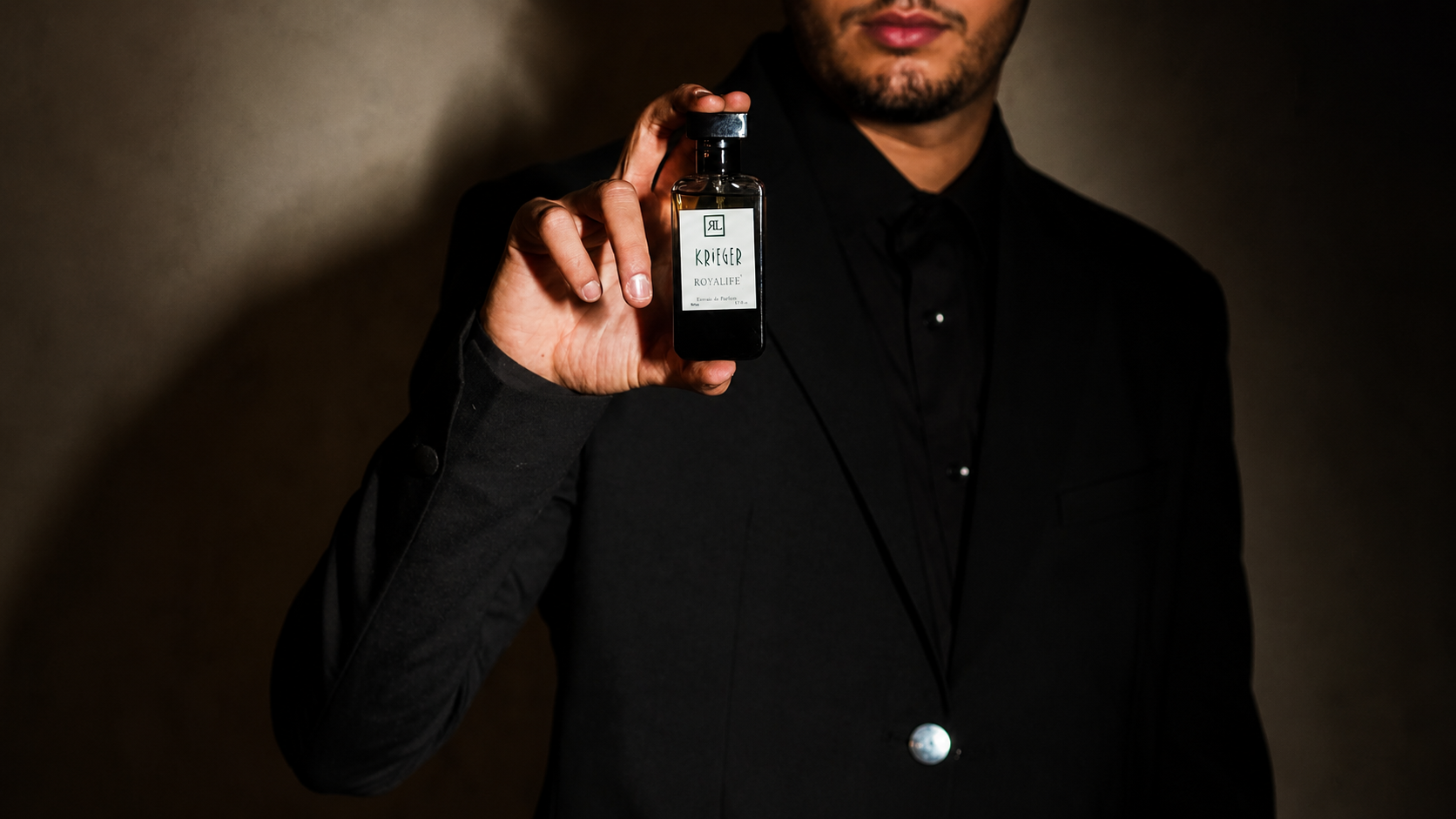 A man in a black suit holds up a small dark glass bottle with a white label towards the camera against a dark brown background.